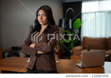 A confident Asian businesswoman stands with her arms crossed in her office, looking at the camera. 116981498