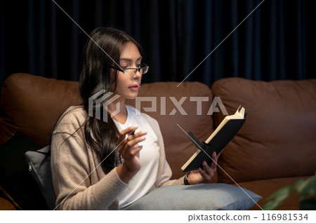 A beautiful Asian woman wearing eyeglasses is reading a book, writing in her planner on a sofa. A beautiful Asian woman wearing eyeglasses is reading a book, writing in her planner on a sofa. 116981534
