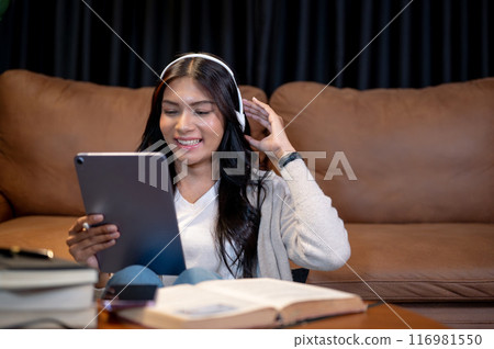 A happy Asian woman is smiling at her tablet, using her digital tablet in a living room at night. A happy Asian woman is smiling at her tablet, using her digital tablet in a living room at night. 116981550