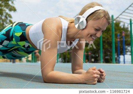 Strong athletic woman with headphones doing exercises on sports field, close up Strong athletic woman with headphones doing exercises on sports field, close up 116981724