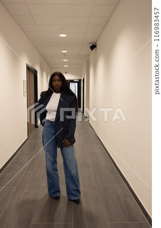 Confident young Black woman in elegant blazer and jeans walking in modern office corridor Confident young Black woman in elegant blazer and jeans walking in modern office corridor 116983457