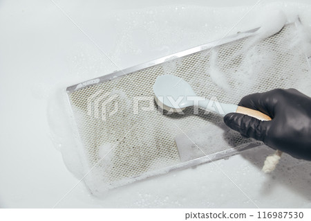 Hand in black glove cleaning mesh filter of cooker hood with brush and foam, white background Hand in black glove cleaning mesh filter of cooker hood with brush and foam, white background 116987530