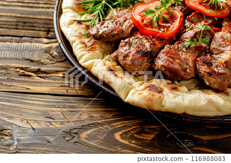 pieces of meat fried on a spit served on thick pita bread with the addition of herbs and vegetables, close-up on a dish on an old dark rough wooden table pieces of meat fried on a spit served on thick pita bread with the addition of herbs and vegetables, close-up on a dish on an old dark rough wooden table 116988083