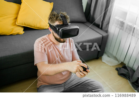 Cheerful young man seated on a sofa playing video games at home. Gaming addiction and entertainment Cheerful young man seated on a sofa playing video games at home. Gaming addiction and entertainment 116988489