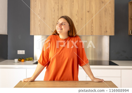 Portrait of smiling young housewife standing near counter in modern kitchen. Home concept. Copy space. 30s Woman dreaming at new home Portrait of smiling young housewife standing near counter in modern kitchen. Home concept. Copy space. 30s Woman dreaming at new home 116988994