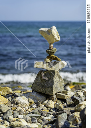 Balanced stone tower stands precariously on rocky beach, with blue ocean and foamy waves in the background Balanced stone tower stands precariously on rocky beach, with blue ocean and foamy waves in the background 116989215