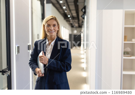 Confident businesswoman holding laptop while standing in modern office hall Confident businesswoman holding laptop while standing in modern office hall 116989480