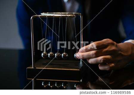 Unrecognizable businessman pulling small metal ball of Newton cradle and observing momentum over glass tabletop Unrecognizable businessman pulling small metal ball of Newton cradle and observing momentum over glass tabletop 116989586