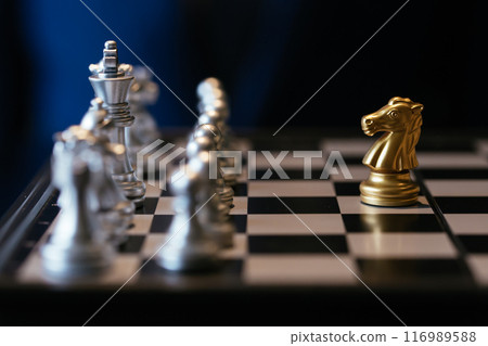Golden knight placed against many silver chess pieces on chessboard with businessman in background Golden knight placed against many silver chess pieces on chessboard with businessman in background 116989588