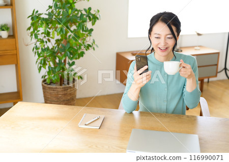A middle-aged woman looking at her smartphone while holding a cup of coffee A middle-aged woman looking at her smartphone while holding a cup of coffee 116990071