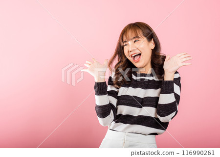 A jubilant woman raises her fists in celebration of her success, saying yes with excitement. Asian portrait of a happy young female in a studio shot on a pink background. 116990261