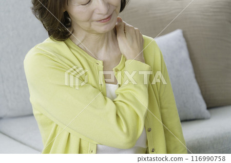 A middle-aged woman sitting in the living room holding her shoulders A middle-aged woman sitting in the living room holding her shoulders 116990758