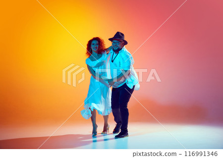 Couple refines their salsa technique, focusing on rhythm and fluidity, showcasing their passion for Latin dance in neon light against gradient orange background. Couple refines their salsa technique, focusing on rhythm and fluidity, showcasing their passion for Latin dance in neon light against gradient orange background. 116991346