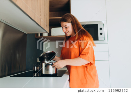 Young happy woman cooking at the kitchen, girl standing near stove at home. Domestic life and leisure moments. High quality photo. Beautiful female preparing tasty meal. 116992552