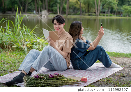 Young Couple Enjoying a Relaxing Picnic by the Lake with Books and Flowers in a Serene Park Setting Young Couple Enjoying a Relaxing Picnic by the Lake with Books and Flowers in a Serene Park Setting 116992597