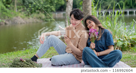 Romantic Couple Enjoying a Relaxing Picnic by the Lake with Flowers and a Book in a Serene Natural Setting Romantic Couple Enjoying a Relaxing Picnic by the Lake with Flowers and a Book in a Serene Natural Setting 116992604