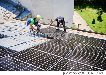 Workers building solar panel system on metal rooftop of house. Two men installers installing photovoltaic solar module outdoors. Alternative, green and renewable energy generation concept. 116993421