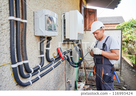 Man electrician installing solar panel system. Technician in helmet and gloves making electrical wiring inverter and electric box. Concept of alternative and renewable energy. Man electrician installing solar panel system. Technician in helmet and gloves making electrical wiring inverter and electric box. Concept of alternative and renewable energy. 116993424