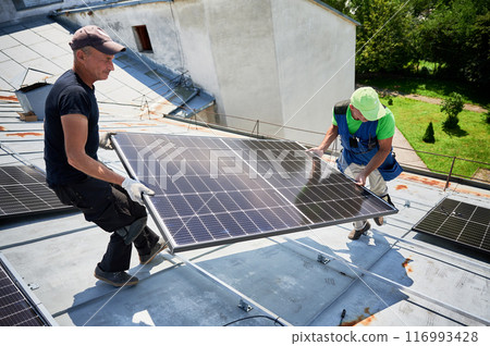 Workers building solar panel system on metal rooftop of house. Two men installers carrying photovoltaic solar module outdoors. Alternative, green and renewable energy generation concept. 116993428