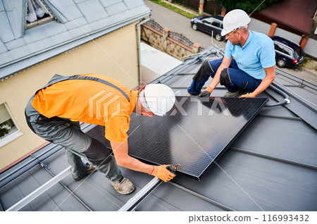 Workers building solar panel system on rooftop of house. Two men installers in helmets installing photovoltaic solar module outdoors. Alternative, green and renewable energy generation concept. 116993432