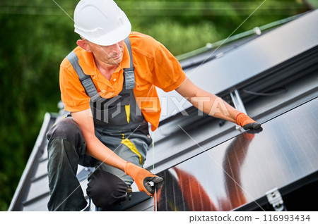 Worker building solar panel system on rooftop of house. Man engineer in helmet installing photovoltaic solar module outdoors. Alternative, green and renewable energy generation concept. 116993434