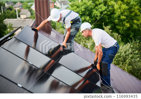 Workers building solar panel system on rooftop of house. Two men installers in helmets installing photovoltaic solar module outdoors. Alternative, green and renewable energy generation concept. Workers building solar panel system on rooftop of house. Two men installers in helmets installing photovoltaic solar module outdoors. Alternative, green and renewable energy generation concept. 116993435