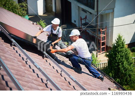 Workers building solar panel system on rooftop of house. Two men installers in helmets installing photovoltaic solar module outdoors. Alternative, green and renewable energy generation concept. Workers building solar panel system on rooftop of house. Two men installers in helmets installing photovoltaic solar module outdoors. Alternative, green and renewable energy generation concept. 116993436