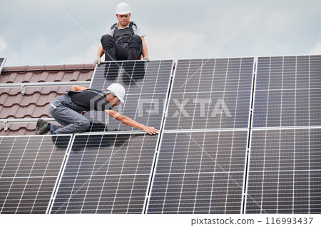 Men mounters mounting photovoltaic solar moduls on roof of house. Electricians in helmets installing solar panel system outdoors. Concept of alternative and renewable energy. Men mounters mounting photovoltaic solar moduls on roof of house. Electricians in helmets installing solar panel system outdoors. Concept of alternative and renewable energy. 116993437