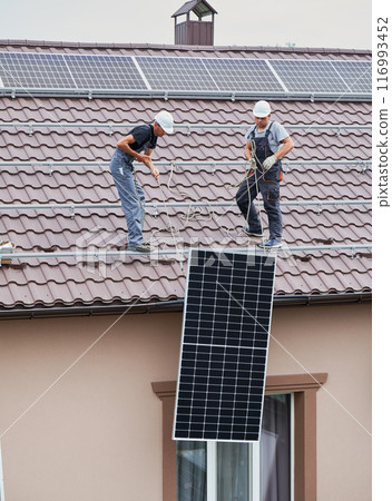 Men installers lifting up photovoltaic solar moduls on roof of house. Electricians in helmets building solar panel system outdoors. Concept of alternative and renewable energy. Men installers lifting up photovoltaic solar moduls on roof of house. Electricians in helmets building solar panel system outdoors. Concept of alternative and renewable energy. 116993452