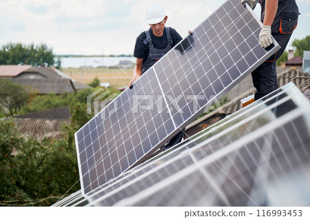 Men technicians mounting photovoltaic solar moduls on roof of house. Builders in helmets installing solar panel system outdoors. Concept of alternative and renewable energy. Men technicians mounting photovoltaic solar moduls on roof of house. Builders in helmets installing solar panel system outdoors. Concept of alternative and renewable energy. 116993453