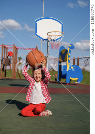 A cheerful playful girl in a Checkered shirt with a basketball is sitting on sports court A cheerful playful girl in a Checkered shirt with a basketball is sitting on sports court 116993776