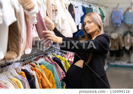 Pregnant woman choosing dress and bodysuits at clothing store. Mother doing shopping in baby shop. Baby clothes sale and pregnancy concept. 116994116