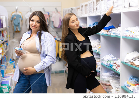 Two pregnant friends shopping together in clothes store. Mothers doing shopping in baby shop. Baby clothes sale and pregnancy concept. Two pregnant friends shopping together in clothes store. Mothers doing shopping in baby shop. Baby clothes sale and pregnancy concept. 116994133