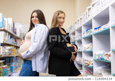 Two pregnant friends shopping together in clothes store. Mothers doing shopping in baby shop. Baby clothes sale and pregnancy concept. Two pregnant friends shopping together in clothes store. Mothers doing shopping in baby shop. Baby clothes sale and pregnancy concept. 116994134