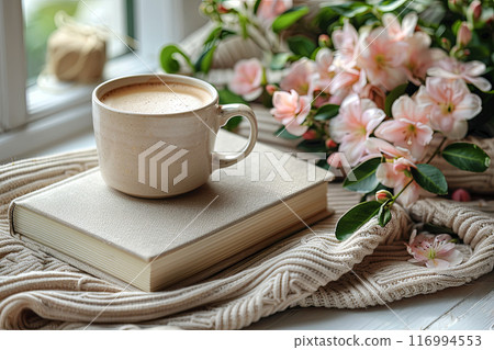 Beige mug with coffee sits on top of a book and pink flowers near windows. Concept of relaxation and leisure Beige mug with coffee sits on top of a book and pink flowers near windows. Concept of relaxation and leisure 116994553
