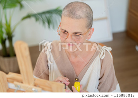 Senior Woman with Shaved Head Painting in a Bright, Modern Studio with Natural Light 116995012