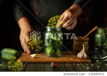 The cook adds dill to a jar of cucumbers before canning. The concept of making pickled delicious cucumbers on the kitchen table by the hands of the chef The cook adds dill to a jar of cucumbers before canning. The concept of making pickled delicious cucumbers on the kitchen table by the hands of the chef 116996542