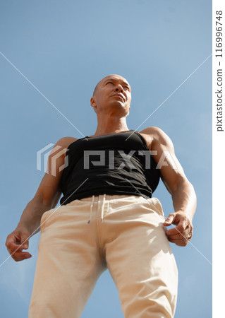 Strict sports coach in black looking away against blue sky. Portrait of athletes man or fitness trainer. Summer. Outdoors. Hero view. Vertical. 116996748