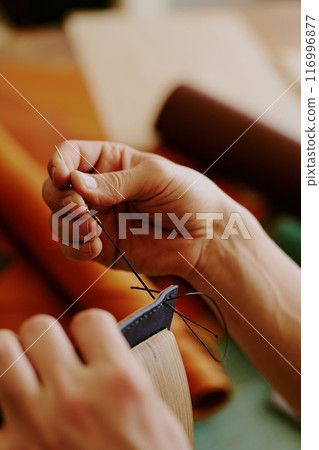 Unrecognizable male hands holding huge needle with black thread to sew leather workpiece 116996877