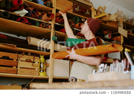 Low angle shot of male tailor taking several leather rolls from wooden shelves 116996888