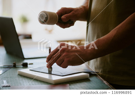 Medium close up of strong male hands using leather punch to poke leather workpiece 116996891