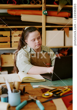 Female atelier worker with down syndrome typing on laptop in old atelier with wooden shelves 116996893