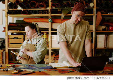 Male tailor using black laptop to check online orders while his female coworker with down syndrome using digital, tablet 116996897