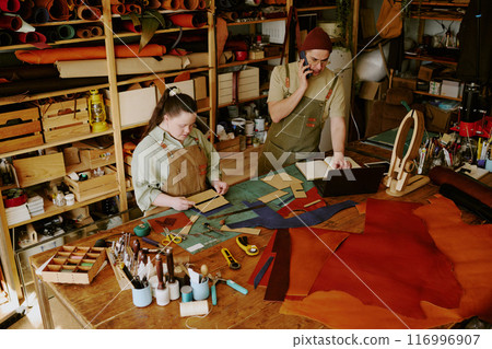 Female tailor with down syndrome cutting out workpieces while her male colleague taking orders via phone 116996907
