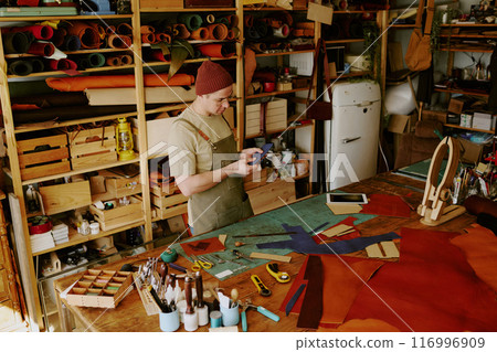 High angle shot of male tailor holding deep blue colored leather workpiece and looking at it at work in fully equipped atelier 116996909