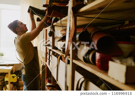 Male tailor sorting multicolored leather rolls on shelves in old atelier 116996918