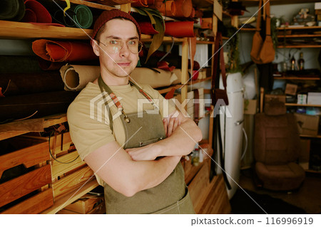 Male tailor with crossed arms smiling lightly and leaning on material storage while posing for portrait 116996919