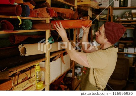 Male tailor taking caramel colored leather roll from upper shelf in atelier Male tailor taking caramel colored leather roll from upper shelf in atelier 116996920