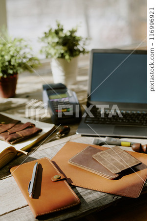 Handmade leather notebooks and card holders on old wooden table, laptop and payment terminal in background 116996921