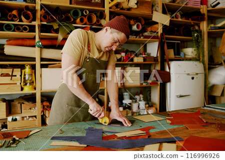 Focused Caucasian tailor working with deep blue colored leather fabric to sew leather goods in atelier 116996926